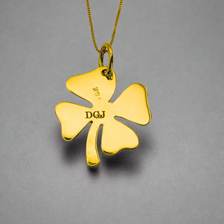 A close-up image of a gold clover leaf pendant with the DGJ (Delma Gold Jewelry) logo engraved on the back. The pendant is attached to a fine gold chain, showcasing its polished finish and intricate design. The engraving of the DGJ logo adds a touch of authenticity and brand recognition, making this four-leaf clover pendant not just a symbol of luck but also a statement of style and quality craftsmanship.