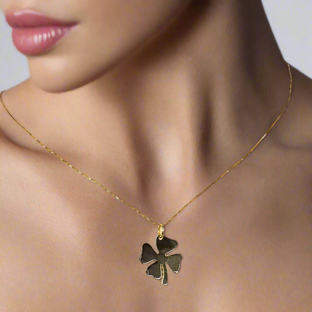 A close-up image of a woman wearing a delicate gold necklace featuring a clover leaf pendant. The pendant sits elegantly on her neckline, showcasing its shiny, polished finish. The simple yet sophisticated design highlights the elegance and charm of the jewelry, making it a standout accessory for everyday wear or special occasions.